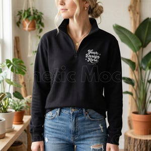Puede incluir: Una sudadera negra con cremallera de cuarto de cierre con el texto "Your Design Here" en blanco. La sudadera tiene un cuello alto y mangas largas. La modelo lleva vaqueros azules. El fondo incluye plantas y una superficie de madera.