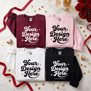 Op de afbeelding: Vier crewneck sweatshirts in bordeaux, roze, wit en zwart, elk met de tekst "Your Design Here" in een retro lettertype. De sweatshirts zijn gerangschikt op een marmeren oppervlak met decoratieve elementen.