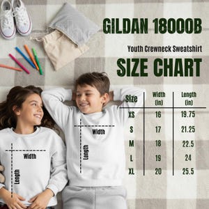 Puede incluir: Dos niños con sudaderas blancas de cuello redondo están acostados sobre una manta a cuadros. La imagen incluye una tabla de tallas para sudaderas juveniles, con medidas en pulgadas. También se ve el texto "GILDAN 18000B" y "Youth Crewneck Sweatshirt".