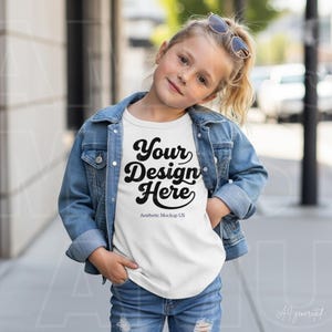 Op de afbeelding: Een wit t-shirt met de zwarte tekst Your Design Here en Aesthetic Mockup US, gedragen onder een blauw spijkerjack. Het kledingstuk wordt gecombineerd met een blauwe spijkerbroek en bevat een zonnebril die boven op het hoofd rust.