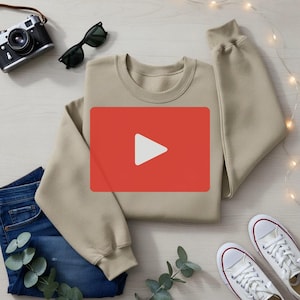 May include: A beige crewneck sweatshirt with a red rectangle overlaying the center, featuring a white play button. The sweatshirt is paired with blue jeans, white sneakers, a vintage camera, and sunglasses. Decorative lights and greenery complete the casual ensemble.