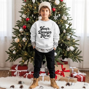 May include: A young person wearing a white sweatshirt with the text "Your Design Here" in black script. They are wearing a beige knit cap, gray long-sleeve shirt, black cargo pants, and tan boots. A decorated Christmas tree is in the background.