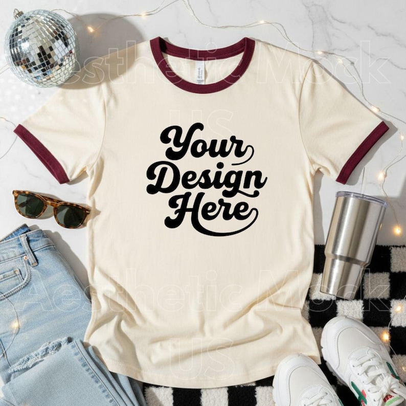 May include: Cream ringer t-shirt with burgundy trim and the text "Your Design Here" in black. The image includes sunglasses, jeans, a stainless steel tumbler, a disco ball, and white trainers.