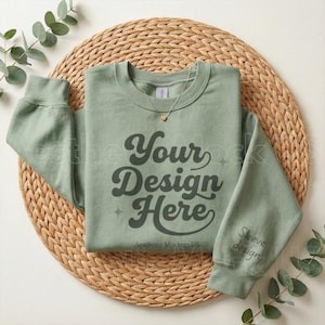 May include: Sage green sweatshirt with the text "Your Design Here" in a retro font. The sleeve has the text "Sleeve design". A gold necklace with a heart pendant is on the neckline. The sweatshirt is on a woven, circular placemat.