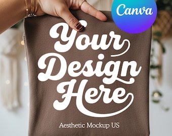 May include: A brown t-shirt featuring the white text Your Design Here in a bold, cursive font. The shirt is held up against a blurred background, and a circular blue and purple Canva logo is visible in the upper right corner of the image.