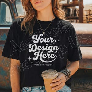 Op de afbeelding: Zwart T-shirt met de tekst "Your Design Here" in een witte, retro lettertype. De persoon draagt een doorzichtige plastic beker met een bruine drank. De persoon draagt armbanden en een ketting.