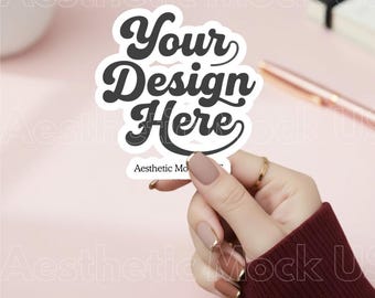 May include: A white die-cut sticker featuring the bold black text Your Design Here in a retro script font, with the smaller text Aesthetic Mockups printed below. The sticker is held up against a soft pink background with a pen and notebook.