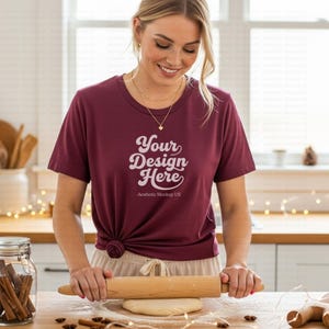 May include: A burgundy t-shirt with the text "Your Design Here" is worn by a person rolling dough with a wooden rolling pin. The text "Aesthetic Mockup US" is below the main text. The scene is set in a kitchen.
