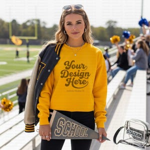 May include: A mustard yellow crewneck sweatshirt featuring the text Your Design Here in dark brown script, worn with a dark grey varsity jacket draped over one shoulder and holding a dark grey felt pennant with the word SCHOOL in white.