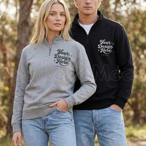 May include: A gray quarter-zip sweatshirt and a black quarter-zip sweatshirt. Both sweatshirts have the text "Your Design + Here" printed on the front. The models are wearing blue jeans. The background is a blurred outdoor scene.