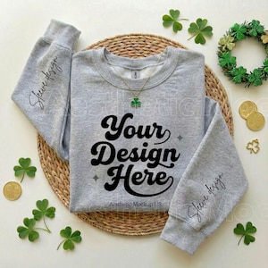 May include: A heather gray sweatshirt with the text "Your Design Here" in a bold, retro font. The sleeves have the text "Sleeve design". A gold necklace with a green shamrock pendant is visible. The image is styled with shamrocks and gold coins.