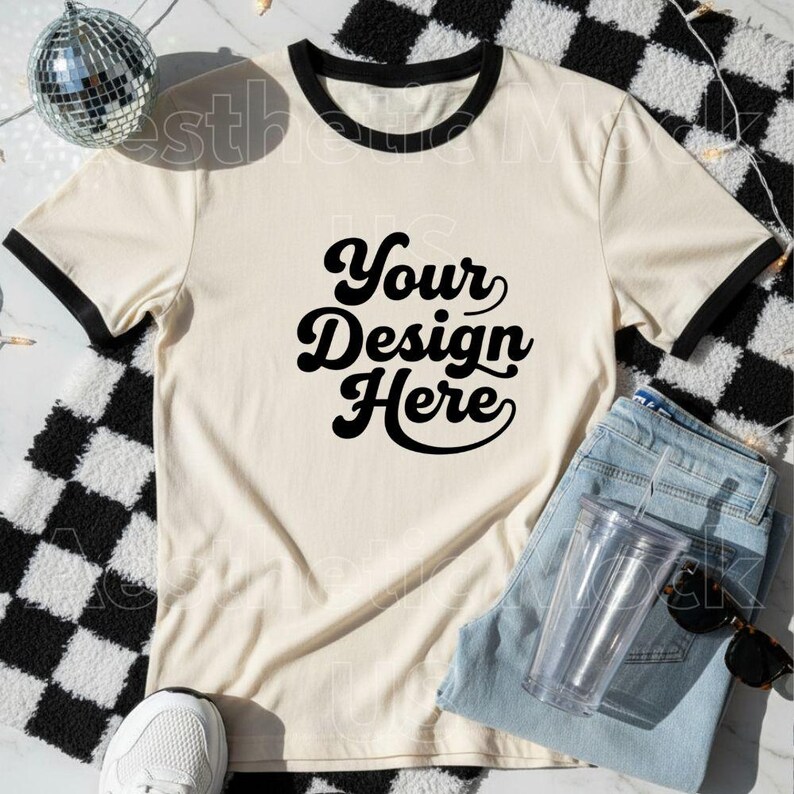 May include: A cream-coloured t-shirt with black trim and the text "Your Design Here" in a bold, retro font. The shirt is paired with light-wash denim jeans, a clear tumbler, sunglasses, and a white trainer. A disco ball and a black and white checkered pattern are in the background.