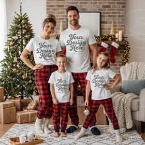 Op de afbeelding: Een gezin van vier personen draagt bijpassende witte t-shirts met de tekst Your Design Here en rood-zwart geruite pyjamabroeken. De groep staat in een woonkamer die is versierd voor de feestdagen met een kerstboom en kerstsokken.