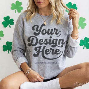 Puede incluir: Una sudadera gris jaspeado de manga larga y cuello redondo con el texto gris oscuro Your Design Here en una fuente cursiva negrita. La sudadera tiene puños en las mangas y se lleva con un collar dorado y dos pulseras de cuentas.