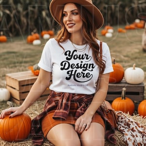 Op de afbeelding: Een wit T-shirt met korte mouwen en de zwarte tekst Your Design Here en Aesthetic Mockup US op de voorkant. Het shirt wordt gecombineerd met een bruin flanellen overhemd met ruitpatroon om de taille, een bruine hoed en een bruine rok.