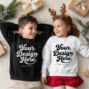 May include: Two children wearing sweatshirts with the text "Your Design Here". One sweatshirt is black, the other is white. The children are lying down, surrounded by wrapped gifts and holiday greenery. The text "Aesthetic Mockup US" is below the main text.