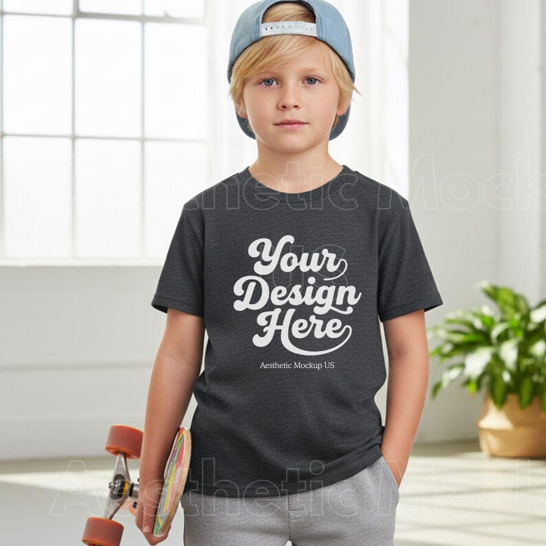 K&ouml;nnte beinhalten: Ein dunkelgraues T-Shirt mit dem Text "Your Design Here" in wei&szlig;er Schrift. Das T-Shirt wird von einer jungen Person getragen, die eine blaue Baseballkappe und eine graue Jogginghose tr&auml;gt. Ein Skateboard wird in einer Hand gehalten.