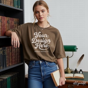 May include: A brown short-sleeve t-shirt featuring the white text Your Design Here and Aesthetic Mockup US on the front. The shirt is paired with blue denim jeans. The garment has a crew neckline and a relaxed fit, shown in a library setting.