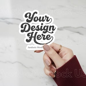 May include: A white sticker with the text "Your Design Here" in a black, retro font. The sticker is held by a hand with a gold ring and maroon sleeve, set against a marble background. The text "Aesthetic Mockup US" is at the bottom.