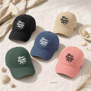 May include: Five baseball caps in black, beige, blue, green, and pink, each featuring the text "Your Design Here" in a black or white script font on the front panel. The caps have a structured crown, a curved brim, and an adjustable back strap.