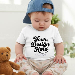 May include: A baby wearing a white t-shirt with the text "Your Design Here" in black script, paired with patterned pants and a blue denim cap. A brown teddy bear is visible on the left side of the image.