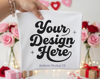 May include: A white square cushion cover featuring the text "Your Design Here" in a bold, black, retro font. The cover is held in front of a blurred background with pink and red roses, gifts, and heart-shaped decorations.