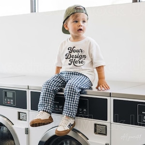 May include: A toddler wearing an olive green baseball cap, a white short-sleeved t-shirt with the black text Your Design Here, and navy blue and white gingham trousers, paired with white and tan trainers, sitting on top of a white laundry machine.