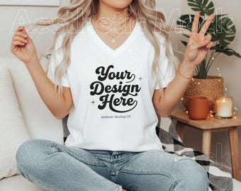 Puede incluir: Camiseta blanca con cuello en V con el texto "Your Design Here" en fuente negra estilizada. La camiseta es parte de una maqueta, y el texto "Aesthetic Mockup US" está debajo. La modelo lleva vaqueros azul claro.