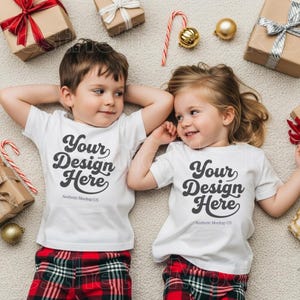 Może przedstawiać: Dwoje dzieci w białych koszulkach z napisem "Your Design Here" i czerwono-kratkowanych spodniach od piżamy. Otaczają je zapakowane prezenty, laski cukrowe i złote ozdoby, sugerujące świąteczny motyw.