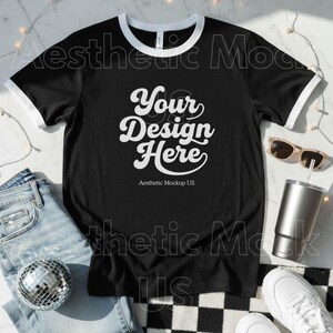 May include: Black ringer t-shirt with white trim, featuring the text "Your Design Here" in a script font. The shirt is displayed with jeans, sunglasses, a tumbler, a disco ball, and white trainers.