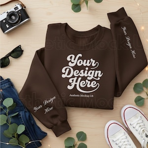 May include: A brown sweatshirt with the text "Your Design Here" in white script. The sleeves have the text "Your Design Here" in white. The sweatshirt is laid out with jeans, sunglasses, a camera, and white sneakers.