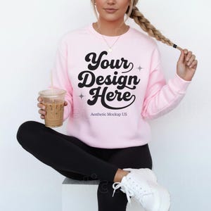 May include: A light pink sweatshirt with the text "Your Design Here" in black script. The sweatshirt is being worn with black leggings and white sneakers. A clear plastic cup with a straw is held in the left hand.