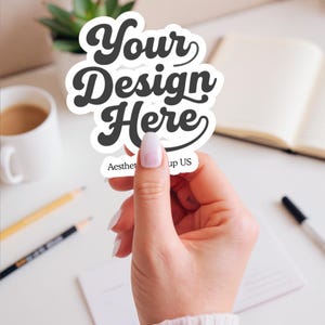 May include: A white sticker with the text "Your Design Here" in a retro font. The sticker is held up by a hand with a manicured nail. The text "Aesthetic Group US" is printed at the bottom of the sticker.