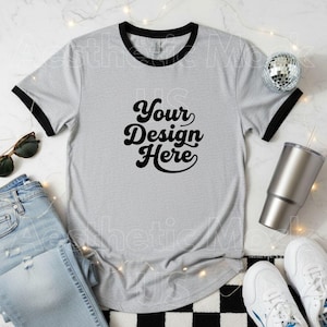 May include: A light grey ringer t-shirt with black trim and the text "Your Design Here" in a black script font. The shirt is displayed with sunglasses, jeans, a disco ball, a silver tumbler, and white trainers.