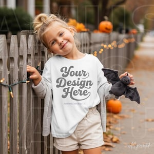 May include: A white t-shirt featuring the gray text Your Design Here and Aesthetic Mockup US, worn with a gray cardigan and beige shorts. A small gray plush bat toy is held in one hand. The shirt displays a clear area for custom graphics.