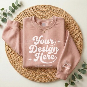 May include: A dusty rose sweatshirt with the text "Your Design Here" in white script. The sleeve has the words "Sleeve design" printed on it. The sweatshirt is on a woven, circular mat with greenery.