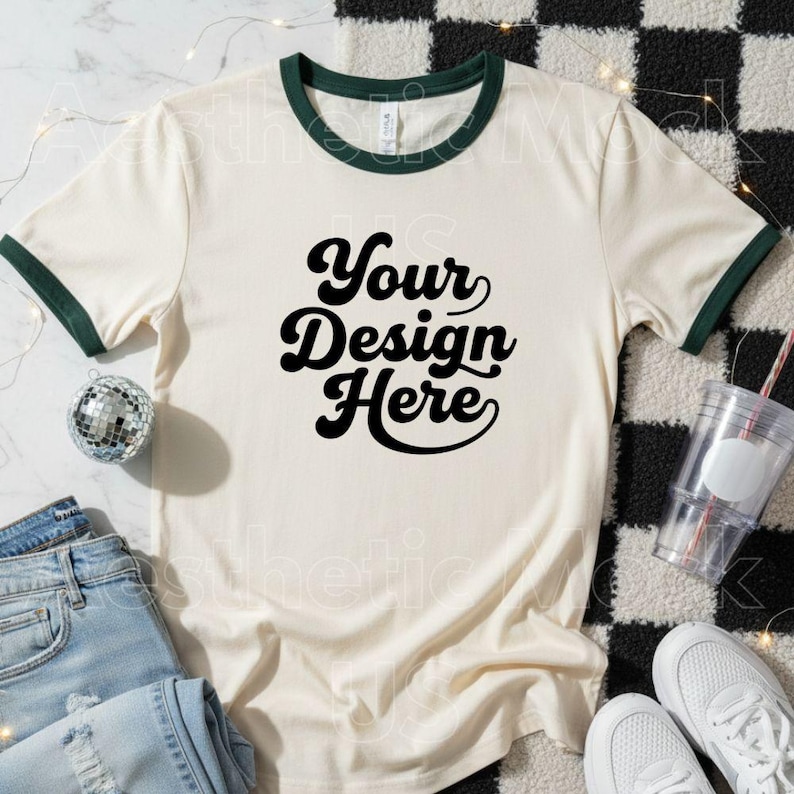 May include: Cream-coloured ringer t-shirt with dark green trim and the text "Your Design Here" in a bold, black font. The shirt is paired with light blue jeans, white trainers, and a disco ball. The background features a black and white checkered pattern.