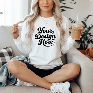 May include: White long-sleeve shirt with the text "Your Design Here" in a black, retro font. The shirt is worn with black shorts and white trainers. The person is holding a clear plastic cup with a straw.