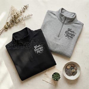 May include: Two quarter-zip sweatshirts, one black and one gray, are displayed on a textured surface. Each sweatshirt features a zipper and the text "Your Design Here." A small bowl with stones and lavender, a succulent, and dried flowers are also present.