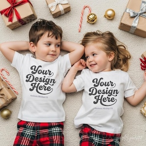 May include: Two white t-shirts with the black text Your Design Here and Aesthetic Mockup US, worn with red and black plaid trousers. The shirts are displayed alongside wrapped gift boxes, a candy cane, and gold ornaments on a textured surface.