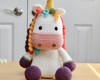 May include: A crocheted unicorn stuffed animal with a white body, pink nose, and purple feet. The unicorn has a yellow horn and a rainbow mane.