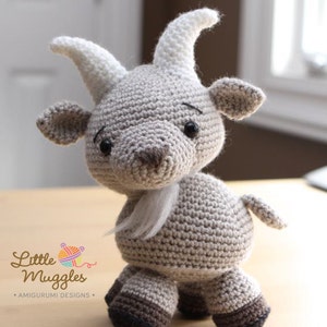 Może przedstawiać: Pluszowa zabawka amigurumi w kształcie kozy, wykonana na szydełku, z brązowo-białym futerkiem, czarnymi oczami i białą brodą. Zabawka stoi na drewnianej powierzchni.