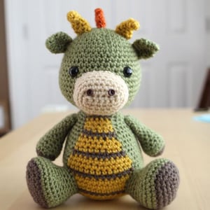 Könnte beinhalten: Ein grünes Häkel-Amigurumi-Spielzeug in Form eines Drachen mit gelben und orangen Hörnern, braunen Füßen und einem gelben und braunen gestreiften Bauch.