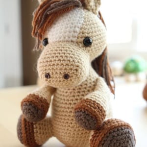 May include: A crocheted brown and white horse amigurumi toy with brown hooves. The toy has a white patch on its face and black eyes.