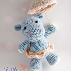 Può includere: Un ippopotamo all'uncinetto blu chiaro con una gonna rosa e gialla tiene un ombrello all'uncinetto grigio chiaro con un bordo rosa. L'ippopotamo ha occhi neri e guance rosa. Lo sfondo è grigio chiaro.