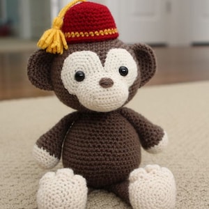 May include: A brown crocheted monkey wearing a red fez with a yellow tassel. The monkey is sitting on a light brown surface.