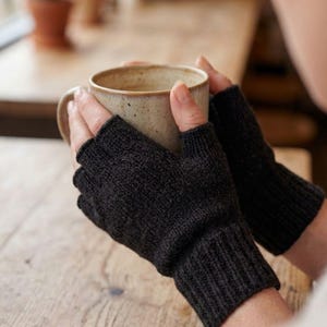Puede incluir: Un par de guantes sin dedos de color gris carbón oscuro con un puño de punto acanalado y un patrón de punto texturizado en la mano. Los guantes se usan mientras se sostiene una taza de cerámica de color beige claro con motas.