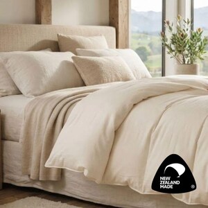 May include: A bed featuring a cream-colored duvet cover, matching pillows, and a textured throw blanket. A black triangular logo in the corner displays the text NEW ZEALAND MADE with a stylized white graphic of a curved shape above the lettering.