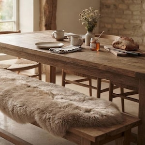 May include: A long, rectangular wooden bench covered with a thick, beige sheepskin rug. The bench is positioned in front of a rustic wooden dining table set with ceramic mugs, a plate, a glass jar of honey, and a loaf of crusty bread.
