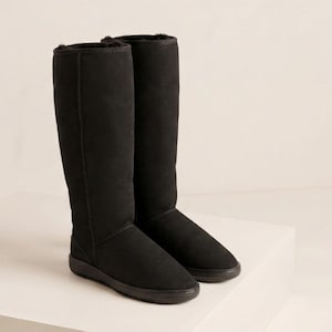 Puede incluir: Un par de botas altas negras con un exterior tipo ante y punta redondeada. Las botas cuentan con una suela negra plana y texturizada, y un forro interior oscuro y afelpado que es visible en la abertura superior de la caña de la bota.